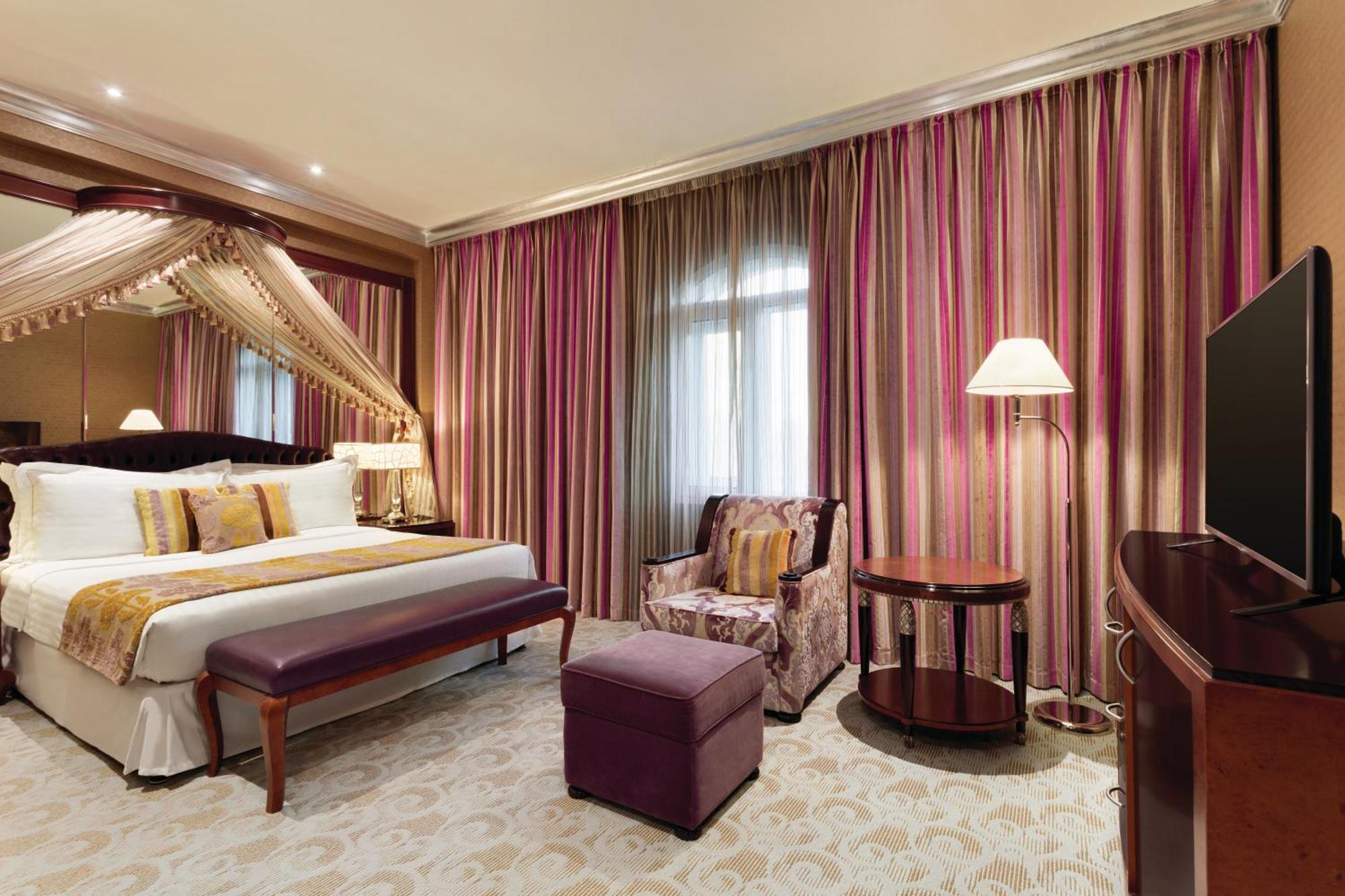 Grand Regency Doha, Trademark Collection By Wyndham Kültér fotó The photo shows a stylish hotel room. It features a large bed with decorative pillows and a draped canopy above it. The room has elegant striped curtains in shades of pink and purple. There is a small seating area with a plush chair and ottoman, alon