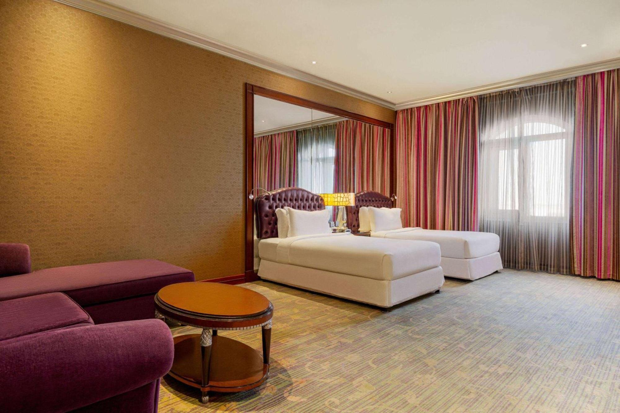 Grand Regency Doha, Trademark Collection By Wyndham Kültér fotó The photo shows a hotel room featuring two beds with white bedding, a large mirror on one wall, and a window dressed with sheer and curtain panels. There is a small round table in the center of the room and a plush purple armchair nearby. The overall